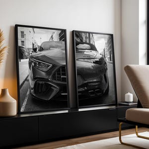 Könnte beinhalten: Zwei gerahmte Schwarz-Weiß-Drucke eines Luxusautos. Die Drucke werden auf einem schwarzen Schrank ausgestellt. Das Auto wird von vorne gezeigt, wobei die Scheinwerfer, der Kühlergrill und die Stoßstange hervorgehoben werden. Der Hintergrund zeigt eine Stadtstraße.