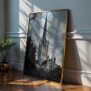 May include: A framed black and white photograph of a cathedral with a tall spire, set against a cloudy sky. The photograph is in a gold frame and leans against a white wall. A potted plant in a gold pot sits nearby on a wooden floor.