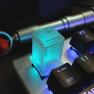 May include: A glowing blue TARDIS keycap on a computer keyboard. The keycap is a miniature replica of the iconic police box. The keyboard keys are black. A silver device is in the background.