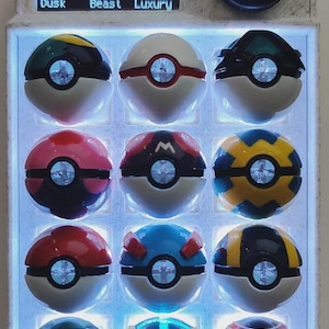 May include: A custom-made PokePad device featuring a screen displaying menu options and a rotary knob. The device showcases a grid of illuminated Poke Balls in various designs. The text "PokePad" is visible on the screen.