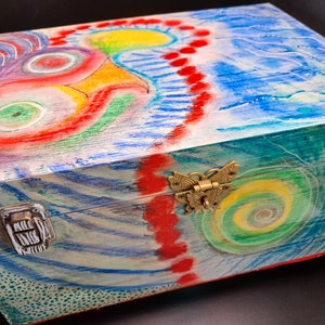 May include: A wooden box with a colorful abstract design painted on the lid. The design features swirls of red, yellow, blue, and green. The box has two brass clasps.