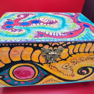 May include: A wooden box with a colorful, abstract design. The box has a butterfly-shaped clasp and is painted in shades of blue, green, yellow, orange, red, and black.
