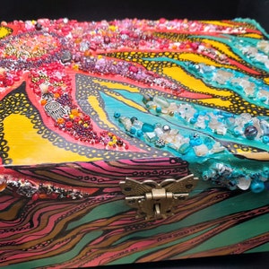 May include: A colorful wooden box with a butterfly latch. The box is decorated with a swirling pattern of red, yellow, blue, and green paint and embellished with beads and other decorative elements.