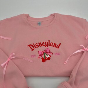 May include: Pink sweatshirt with a red embroidered Disneyland logo, cherries, and a bow. The text "EST. 1955" is also embroidered on the sweatshirt.