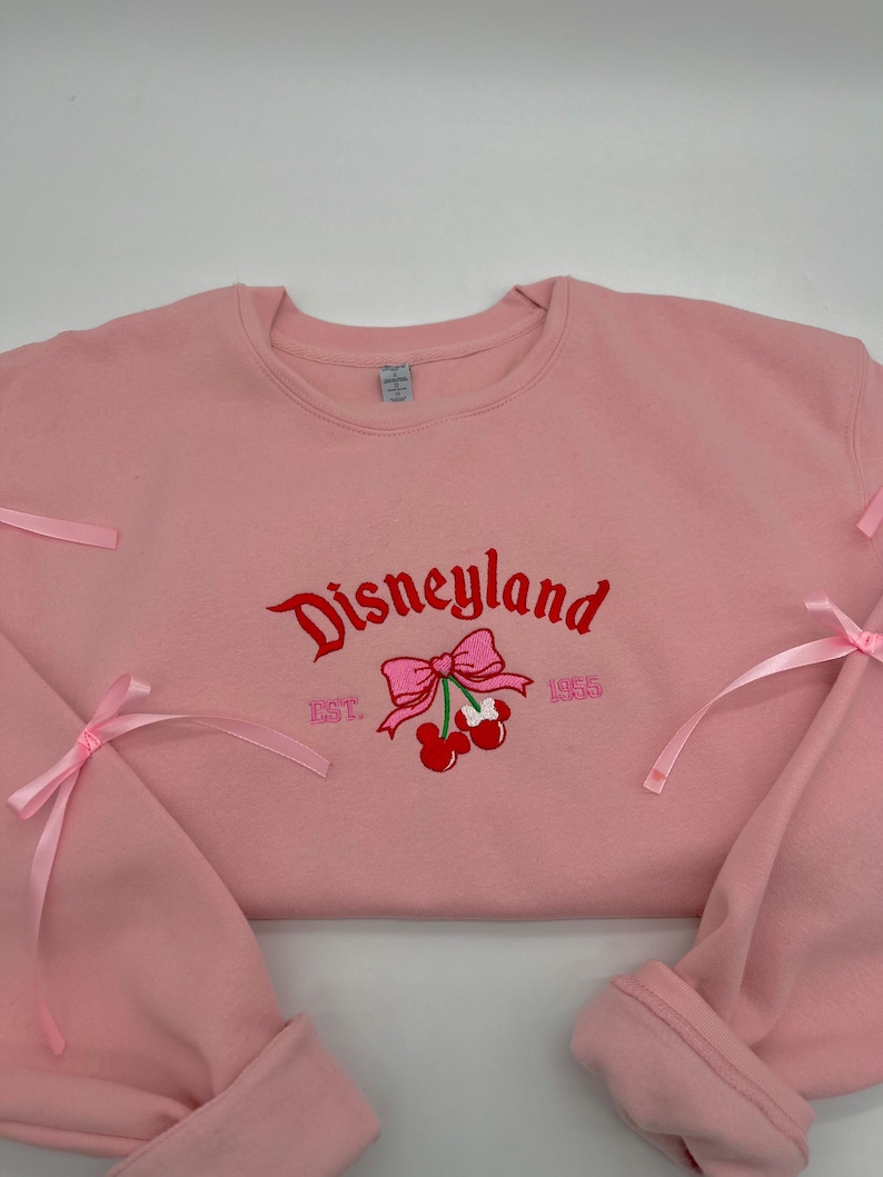May include: Pink sweatshirt with a red embroidered Disneyland logo, cherries, and a bow. The text "EST. 1955" is also embroidered on the sweatshirt.