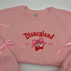 May include: Pink sweatshirt with a red embroidered Disneyland logo, a pink bow with cherries, and the text "EST. 1955".