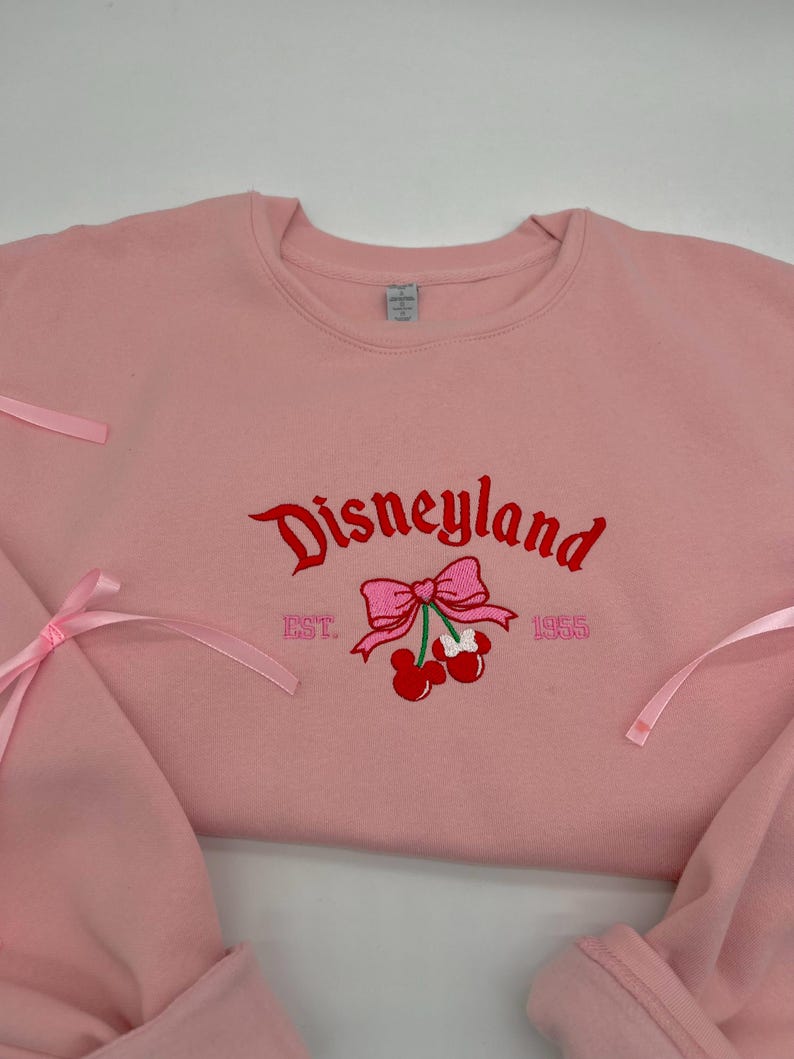 May include: Pink sweatshirt with a red embroidered Disneyland logo, a pink bow with cherries, and the text "EST. 1955".