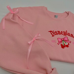 May include: Pink sweatshirt with a red embroidered Disneyland logo and a cherry with a pink bow. The sweatshirt has pink ribbon bows on the sleeves.