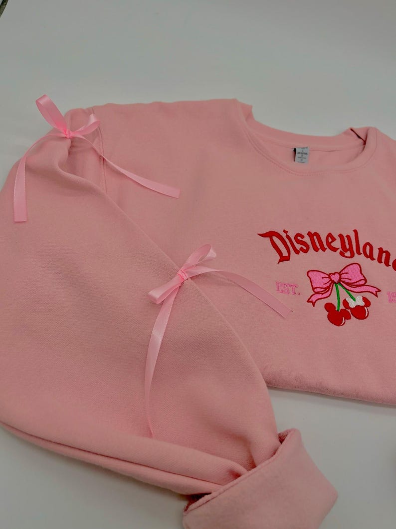 May include: Pink sweatshirt with a red embroidered Disneyland logo and a cherry with a pink bow. The sweatshirt has pink ribbon bows on the sleeves.