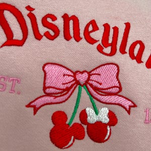 May include: Pink sweatshirt with the text "Disneyland" embroidered in red. A pink bow with a heart in the center is above two red cherries with white Mickey Mouse ears.