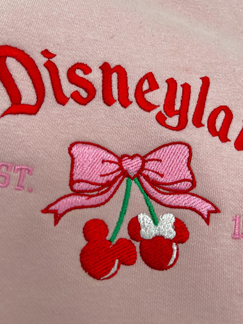 May include: Pink sweatshirt with the text "Disneyland" embroidered in red. A pink bow with a heart in the center is above two red cherries with white Mickey Mouse ears.