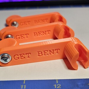 May include: Three bright orange 3D-printed plastic tools arranged in a row. Each tool features a rectangular body with the text GET BENT embossed on the side, a silver metal screw at one end, and a cylindrical slotted opening at the other end.