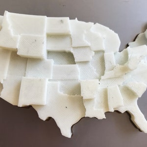 May include: A white 3D printed map of the United States, with each state outlined and raised.