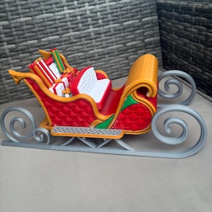 Könnte beinhalten: Ein dekorativer Weihnachtsschlitten in Rot, Gold und Silber, gefüllt mit Geschenken. Der Schlitten hat einen gesteppten roten Körper, goldene Verzierungen und silberne Kufen. Die Geschenke sind in rotes, weißes, grünes und goldenes Papier verpackt.