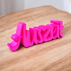 Könnte beinhalten: Das Wort "Auszeit" in leuchtendem Pink, 3D-gedruckt, liegt auf einer Holzoberfläche. Das Wort ist in einer stilisierten Schriftart gehalten, wobei jeder Buchstabe verbunden ist. Der Hintergrund ist eine hellbraune Holzoberfläche.