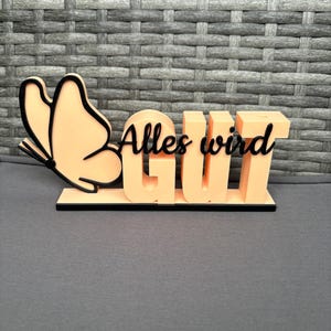 Könnte beinhalten: Ein pfirsichfarbenes Dekoschild mit dem deutschen Satz "Alles wird gut" (Alles wird gut). Das Schild zeigt einen Schmetterling und den Text in Schwarz. Das Schild steht auf einem schwarzen Sockel.