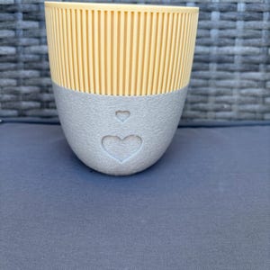 Könnte beinhalten: Ein zweifarbiger Pflanztopf mit einem hellgelben, vertikal gerippten Oberteil und einem grauen, strukturierten Boden. Der graue Bereich weist ein Herzdesign auf. Dieser dekorative Pflanztopf eignet sich für kleine Pflanzen.