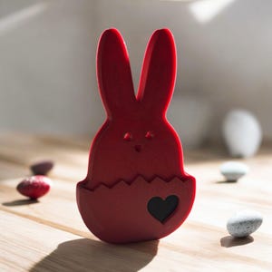 Könnte beinhalten: Eine rot 3D-gedruckte Osterhasenfigur in Form eines Hasen, der aus einem Ei kommt. Der Hase hat einen herzförmigen Ausschnitt auf seiner Brust.