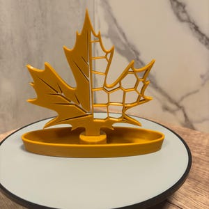 Könnte beinhalten: Eine senfgelbe Ahornblatt-Skulptur. Das Blatt ist geteilt, eine Hälfte massiv und die andere mit einem geometrischen offenen Design. Es sitzt auf einem passenden ovalen Sockel, präsentiert auf einer weißen und schwarzen kreisförmigen Plattform.