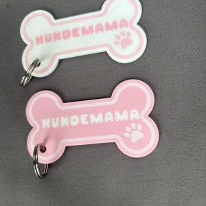 Könnte beinhalten: Zwei knochenförmige Haustieranhänger. Einer ist weiß mit rosa Umrandung und dem Text "HUNDEMAMA" und einem Pfotenabdruck. Der andere ist rosa mit weißer Umrandung, demselben Text und einem Pfotenabdruck. Jeder Anhänger hat einen silbernen Ring.