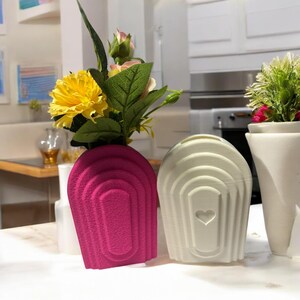 Könnte beinhalten: Zwei Keramikvasen mit einem strukturierten, gewölbten Design. Eine Vase ist pink und die andere ist weiß mit einem Herz-Ausschnitt. Beide Vasen sind mit künstlichen Blumen gefüllt.