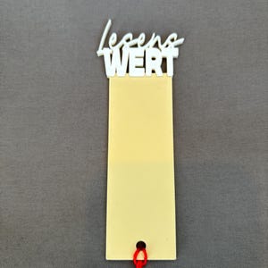 Könnte beinhalten: Ein cremefarbenes Etikett mit den deutschen Worten "LEGEND WERT" in weißer Schrift. Das Etikett hat unten eine rote Schnur. Der Hintergrund ist einfarbig grau.