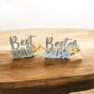 Könnte beinhalten: Zwei dekorative Schilder auf einer Holzoberfläche. Ein Schild zeigt "Best Dad" in grauen Buchstaben mit einem goldenen Hammer und Schraubenschlüssel. Das andere Schild zeigt "Best Papa" in hellblauen Buchstaben mit goldenem Hammer und Schraubenschlüssel.