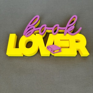 Könnte beinhalten: Ein gelbes "LOVER" Schild mit dem Wort "book" in lila Schrift. Zwischen den Wörtern befindet sich ein kleines lila Buchsymbol. Das Schild ist ein 3D-Rendering, ideal für ein Bücherregal oder einen Schreibtisch. Der Hintergrund ist einfarbig grau.