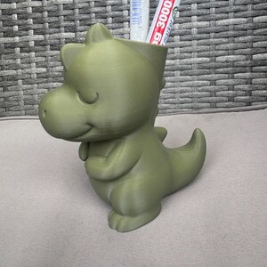 Könnte beinhalten: Ein hellgrüner, 3D-gedruckter Schreibtisch-Organizer in Dinosaurierform. Der Dinosaurier hat einen freundlichen Ausdruck und hält die Arme nah am Körper. Zwei Marker befinden sich im Inneren des Organizers. Die Marker tragen den Text "g 3000 marker".