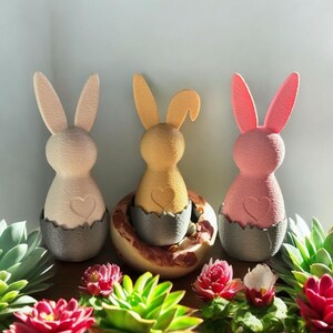 Könnte beinhalten: Drei pastellfarbene Hasenfiguren in eiförmigen Nestern. Die Hasen sind rosa, gelb und weiß. Die Nester sind grau und haben einen gewellten Rand.