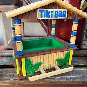 Könnte beinhalten: Eine Miniatur-Tiki-Bar mit einem braunen Holzrahmen, einem beigen Dach und einem grünen Innenraum. Das Schild zeigt "Tiki Bar" in Blau und Weiß. Die Struktur ist mit Bambus-Akzenten und grünen Blattausschnitten verziert.