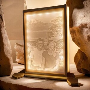 Könnte beinhalten: Ein weißer beleuchteter Bilderrahmen mit einem Foto einer dreiköpfigen Familie, die vor einem See und Bergen steht. Der Rahmen hat einen goldenen Rand und einen Holzsockel.