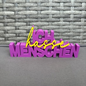 Könnte beinhalten: Ein lila 3D-gedrucktes Schild mit den deutschen Worten "Ich hasse Menschen" in einem Blockbuchstabenstil. Das Wort "hasse" ist in einer gelben Schreibschrift, und das Schild steht vor einem grauen Hintergrund.