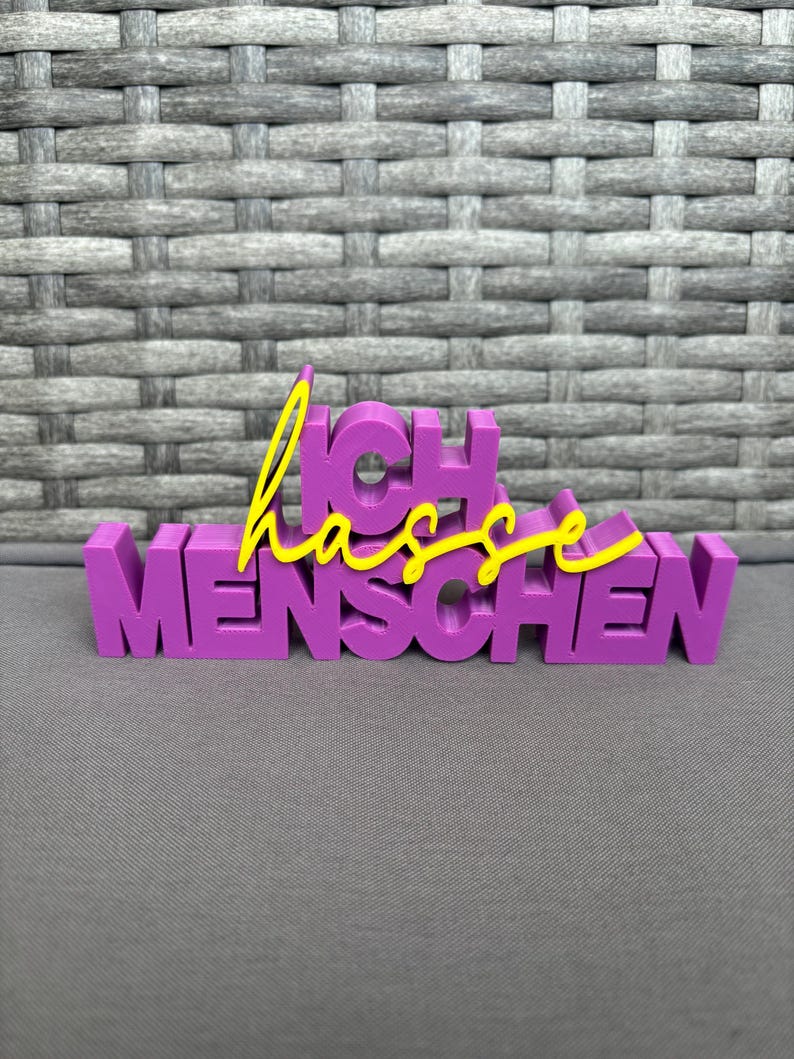 Op de afbeelding: Een paars 3D-geprint bord met de Duitse woorden "Ich hasse Menschen" in een blokletterstijl. Het woord "hasse" staat in een gele cursieve letter, en het bord staat tegen een grijze achtergrond.