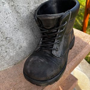 Könnte beinhalten: Ein schwarzer, stiefelförmiger Pflanzkübel mit Schnürdesign. Der Pflanzkübel besteht aus einem dunklen Material und steht auf einer Steinoberfläche. Der Stiefel hat ein strukturiertes Aussehen und eine breite Öffnung zum Bepflanzen.