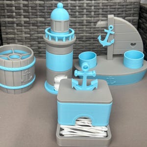 Könnte beinhalten: Ein Badezimmer-Set im maritimen Stil in Grau und Hellblau. Das Set umfasst einen Leuchtturm-förmigen Taschentuchhalter, einen fassförmigen Behälter, einen Bootsförmigen Organizer und einen quadratischen Behälter mit Anker.