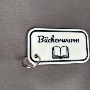 Könnte beinhalten: Ein weißer, rechteckiger Schlüsselanhänger mit schwarzem Rand und silbernem Schlüsselring. Der Schlüsselanhänger zeigt das Wort "Bücherwurm" in schwarzer Schrift über einer schwarzen Illustration eines offenen Buches.