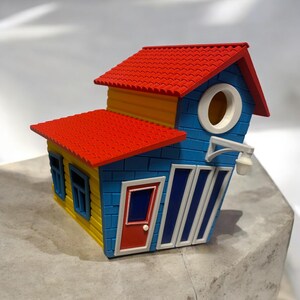 Könnte beinhalten: Ein blaues und gelbes Vogelhaus mit einem roten Dach. Das Vogelhaus hat eine kleine Tür, Fenster und eine Vogelhausöffnung.