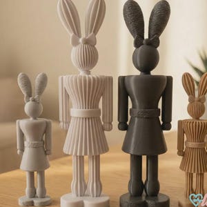 Könnte beinhalten: Vier dekorative Hasenfiguren in verschiedenen Größen und Farben. Die Hasen haben lange Ohren, Schleifen und Kleider. Die Farben sind Weiß, Grau, Schwarz und Hellbraun. Die Figuren werden auf einer Holzoberfläche ausgestellt.