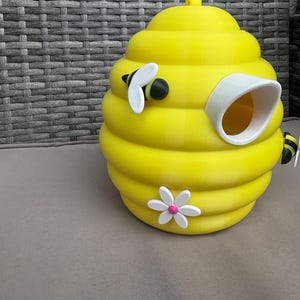 Könnte beinhalten: Ein hellgelbes, bienenstockförmiges Vogelhaus mit einer weißen und rosa Blume und zwei Cartoon-Bienen. Das Vogelhaus hat einen weißen Eingang und einen kleinen gelben Knopf oben. Das Vogelhaus steht auf einer grauen Oberfläche.