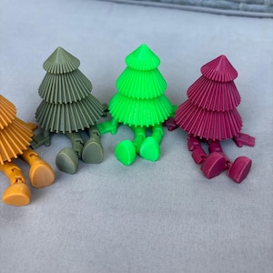 Könnte beinhalten: Eine Sammlung farbenfroher, miniaturisierter Weihnachtsbaumfiguren. Die Bäume sind in den Farbtönen Orange, Grün, Olivgrün und Magenta gehalten. Jeder Baum hat eine konisch geformte Spitze und bewegliche Beine und Füße, was ihm ein verspieltes Aussehen verleiht.