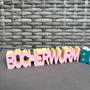 Könnte beinhalten: Rosa und gelbe 3D-gedruckte Buchstaben, die "BÜCHERWURM" buchstabieren, auf einer grauen Oberfläche. Die Buchstaben sind in einer verspielten, blockartigen Schriftart gehalten. Der Hintergrund ist eine graue Webstruktur.