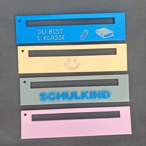Könnte beinhalten: Vier rechteckige, farbige Namensschilder. Das obere Schild ist blau mit dem Text "DU BIST 1. KLASSE" und Bleistift- und Buchsymbolen. Das zweite Schild ist gelb mit einem Smiley. Das dritte Schild ist grau mit dem Wort "SCHULKIND". Das untere Schild ist rosa.