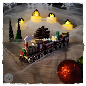 Könnte beinhalten: Ein Miniatur-Weihnachtszug mit drei Waggons, einem rot-grünen Kranz und einer weißen Rauchfahne. Der Zug ist braun, rot und weiß. Die Szene umfasst kleine beleuchtete Häuser, Tannenbäume, einen Tannenzapfen und rote und grüne Christbaumkugeln.