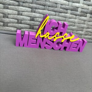 Op de afbeelding: Een paars 3D-geprint bord met de Duitse zin "Ich hasse Menschen" in gele letters. Het bord is in blokletters en staat op een grijze ondergrond.