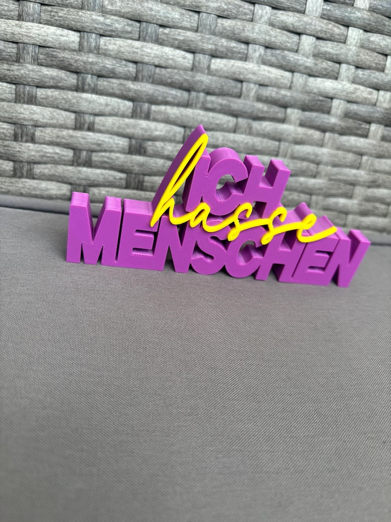 Op de afbeelding: Een paars 3D-geprint bord met de Duitse zin "Ich hasse Menschen" in gele letters. Het bord is in blokletters en staat op een grijze ondergrond.