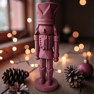 Könnte beinhalten: Eine einfarbige, mauvefarbene Nussknackerfigur mit Krone und Uniform. Die Holzfigur steht auf einem runden Sockel, mit zwei Tannenzapfen und einem dekorativen Ornament in der Nähe. Der Hintergrund umfasst ein Fenster und Lichterketten.