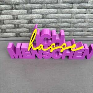 Op de afbeelding: Een paars 3D-geprint bord met de Duitse woorden "Ich hasse Menschen". Het woord "Ich" is in een bloklettertype en het woord "Menschen" is in een vergelijkbaar lettertype. Het woord "hasse" is in een gele cursieve letter.