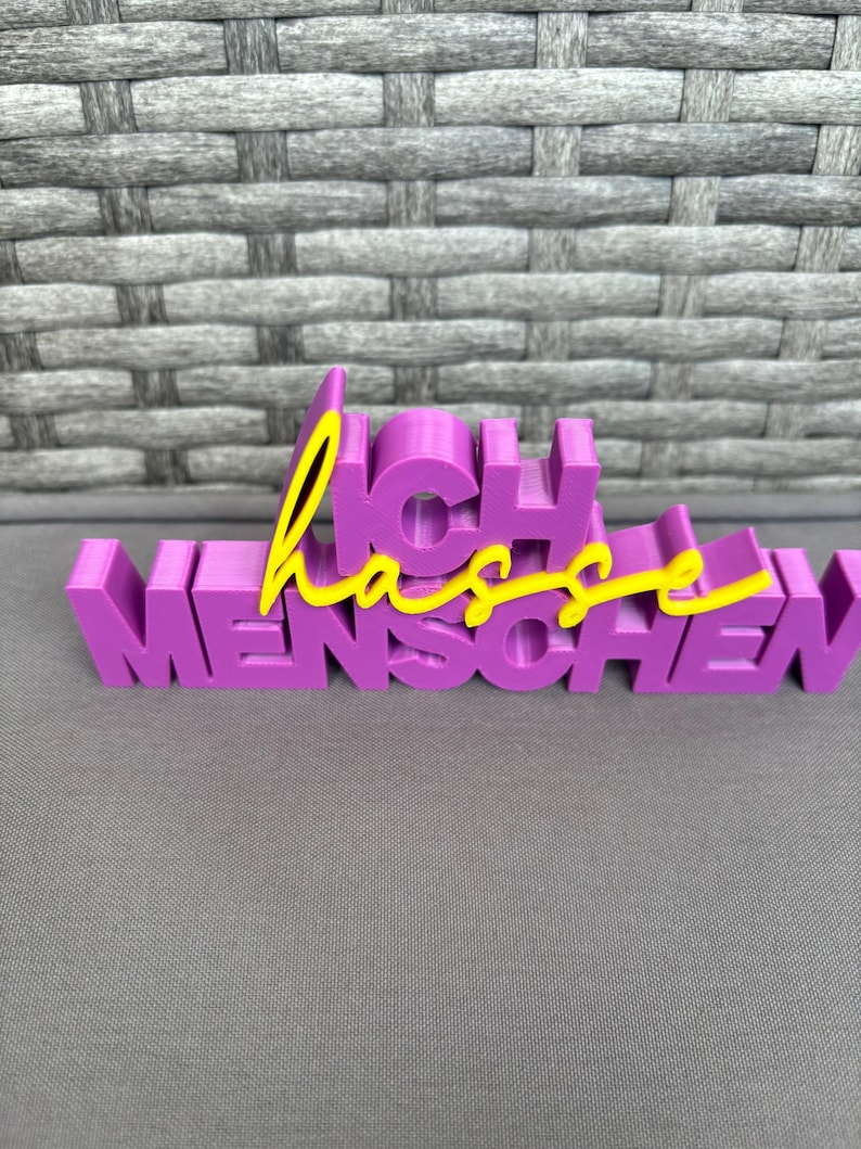 Op de afbeelding: Een paars 3D-geprint bord met de Duitse woorden "Ich hasse Menschen". Het woord "Ich" is in een bloklettertype en het woord "Menschen" is in een vergelijkbaar lettertype. Het woord "hasse" is in een gele cursieve letter.