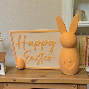 Könnte beinhalten: Osterdekoration in Orange. Eine Hasenfigur mit Herz und ein Schild mit der Aufschrift "Happy Easter". Ein orangefarbenes Ei liegt neben dem Schild. Das Schild hat einen cremefarbenen Hintergrund mit einem orangefarbenen Rand.